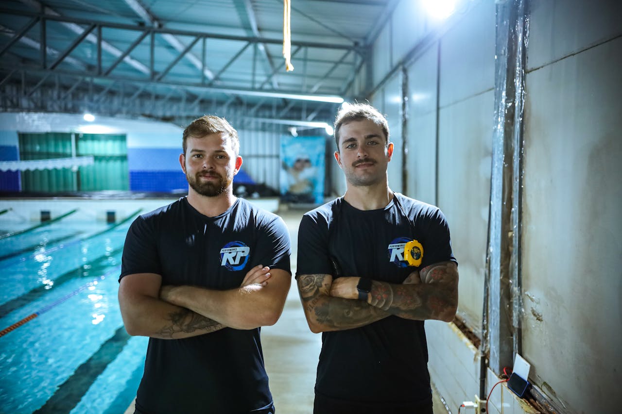Two athletic men standing confidently by an indoor swimming pool, showcasing sports attire.